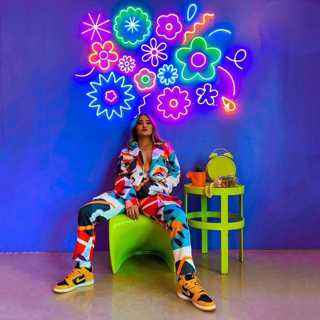 Original cluster of multicolored neon flower and squiggle shapes on a blue wall above a seated person logo featuring blue, neon green, neon pink colours