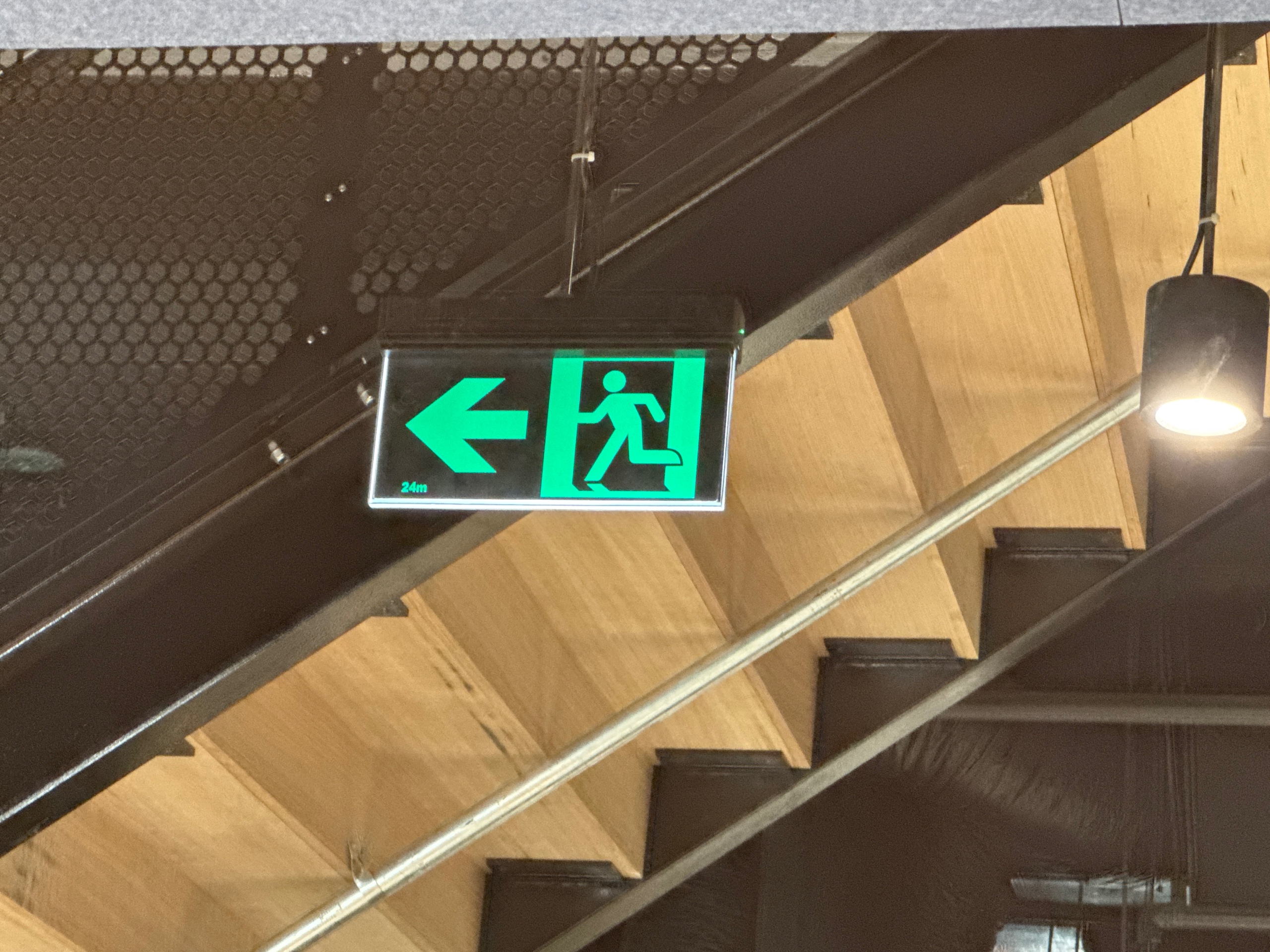 Original green emergency exit sign with a left arrow and running person pictogram on a dark rectangular panel logo featuring green, black, wood-brown colours
