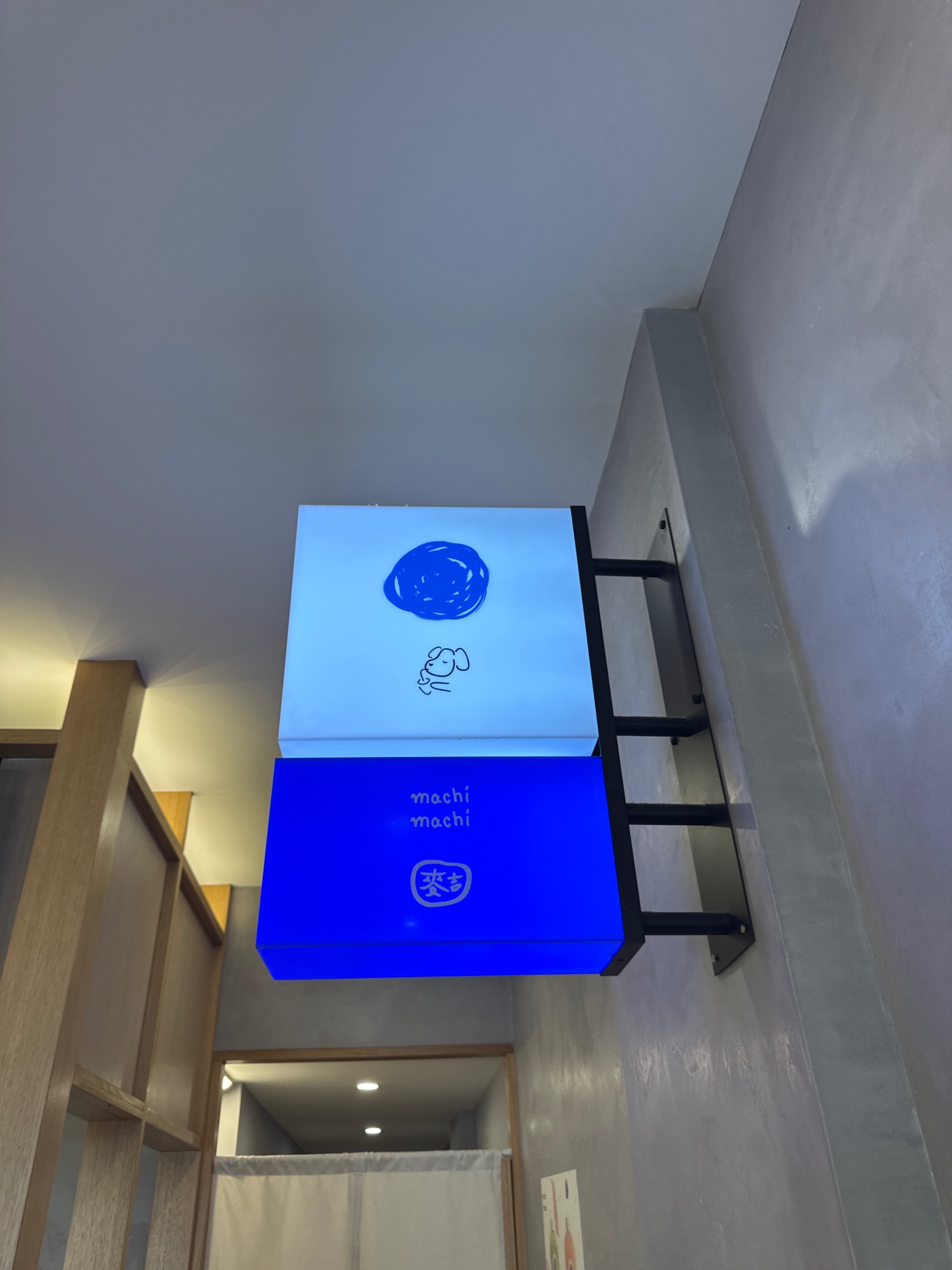 Original white and blue hanging sign with blue circle and dog icon above white text 'machi machi' and a small rounded square mark with white characters logo featuring blue, white, gray colours