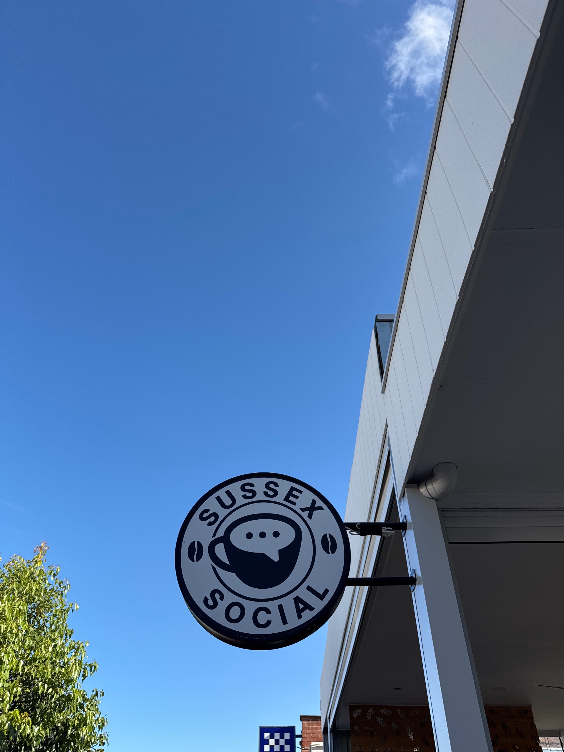 Original circular logo with black coffee cup icon containing a white speech bubble, surrounded by black uppercase text 'SUSSEX' at the top and 'SOCIAL' at the bottom with small coffee bean icons on each side logo featuring blue, white, black colours